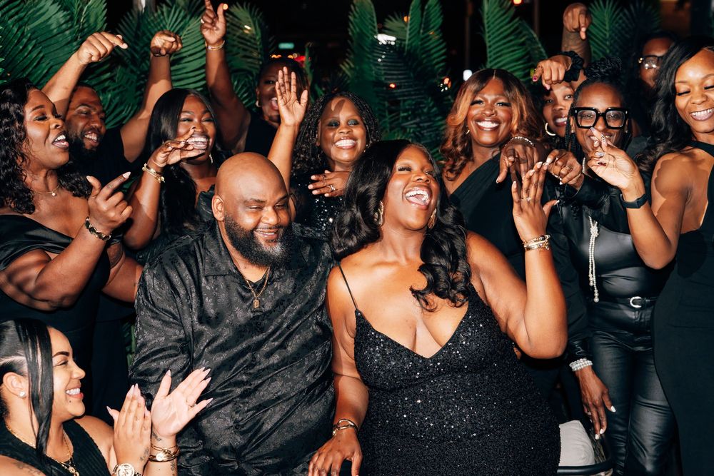 Group of joyful people in elegant black outfits laughing and clapping together at a lively celebration with palm leaves in the background.