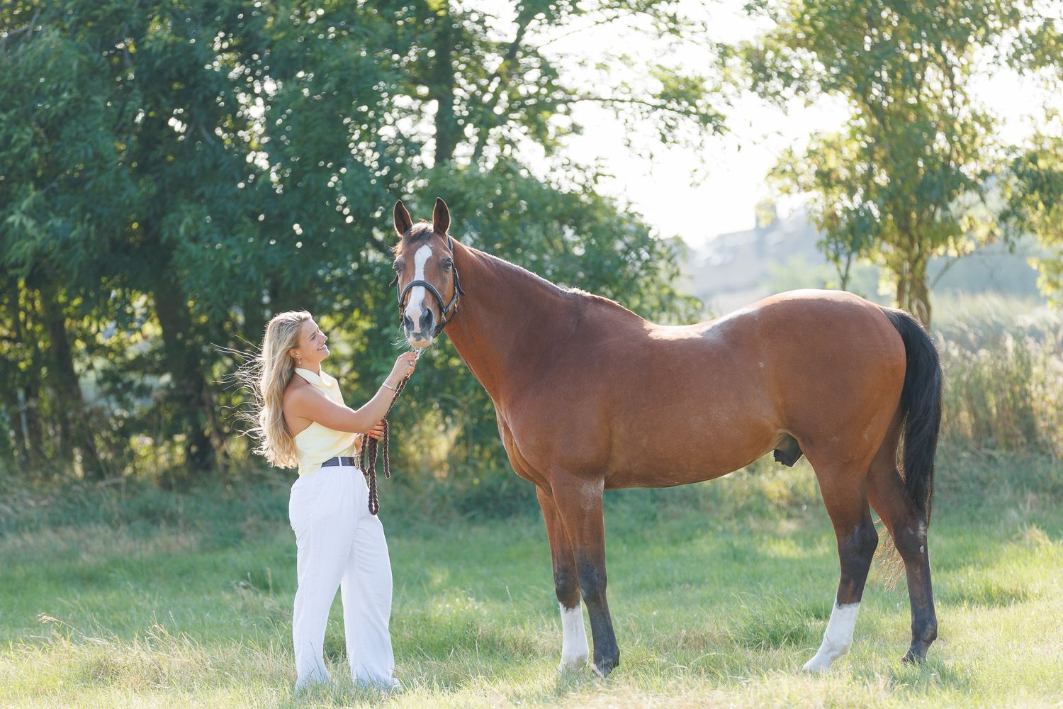 Young woman in black top smiling while gently embracing a brown horse outdoors among lush green trees.