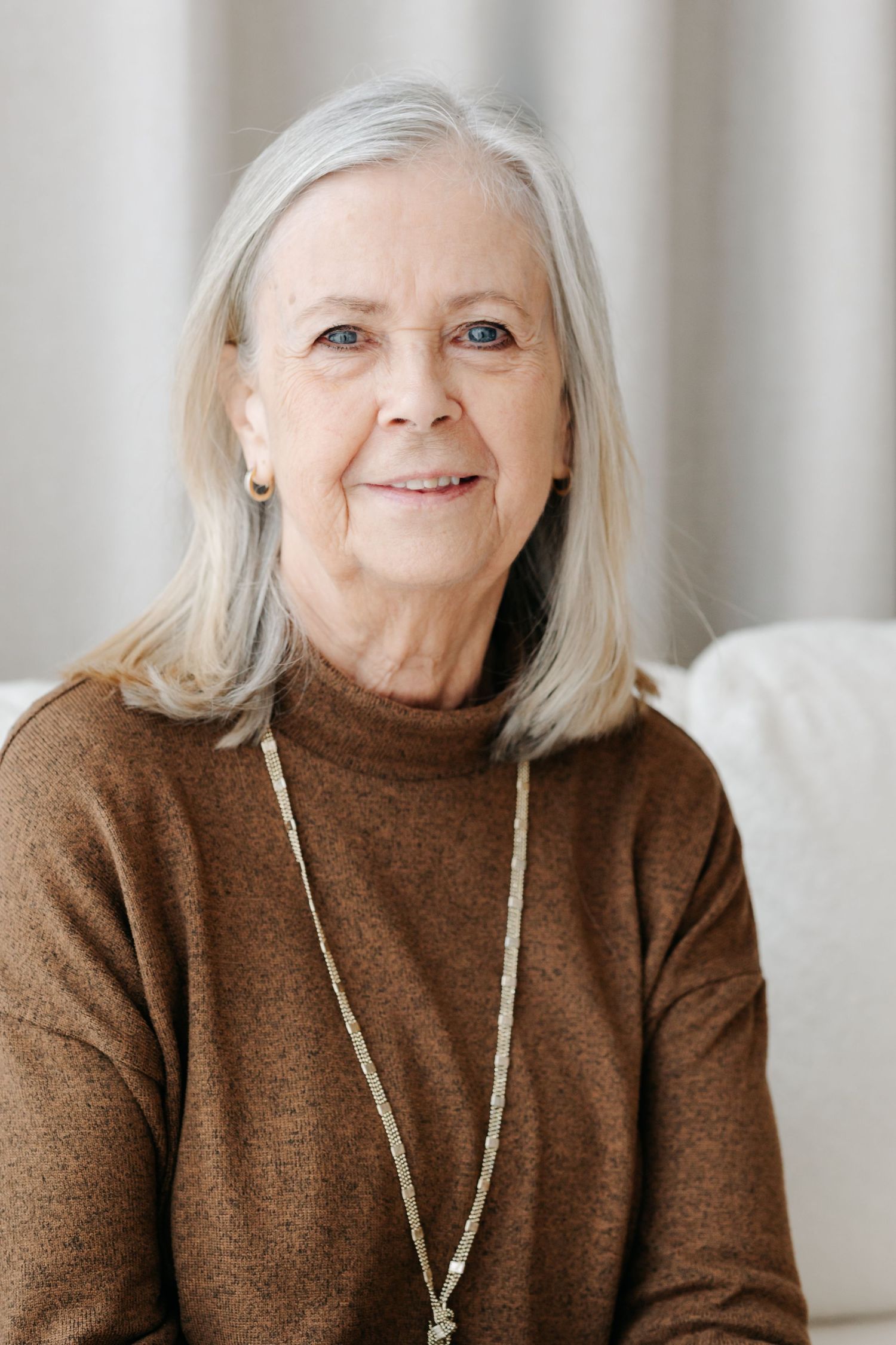 Smiling elderly woman with gray hair wearing a brown turtleneck sweater and long necklace sitting on a white sofa.