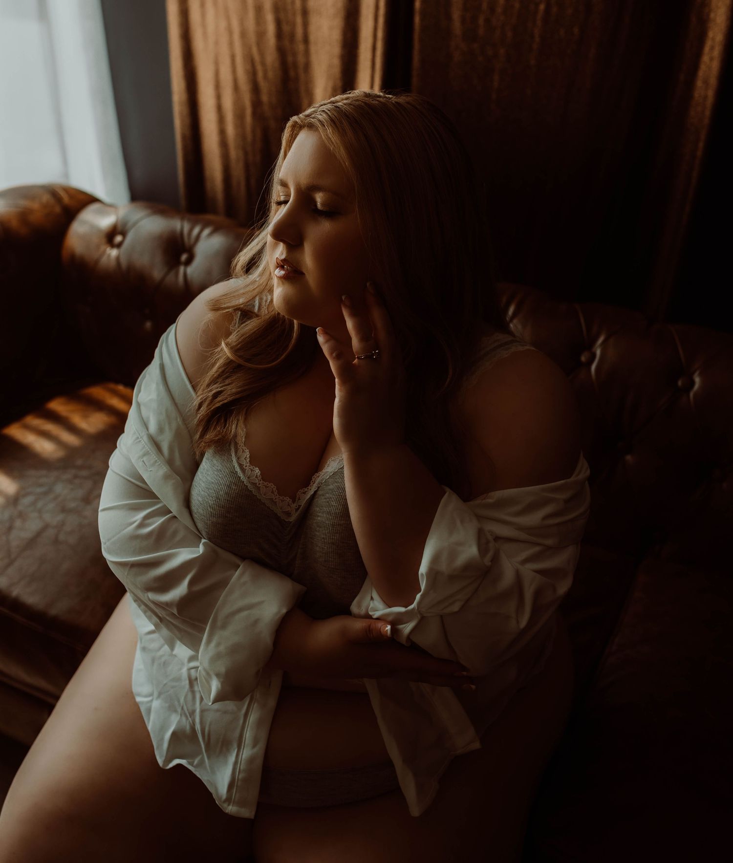 A woman posing in a white shirt and boudoir poses thoughtfully on a leather sofa in warm, moody lighting.