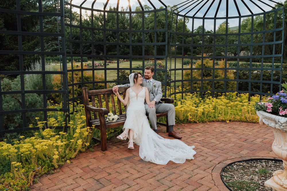 Bride and groom sitting in an elegant iron gazebo surrounded by yellow flowers and lush greenery at a garden wedding.