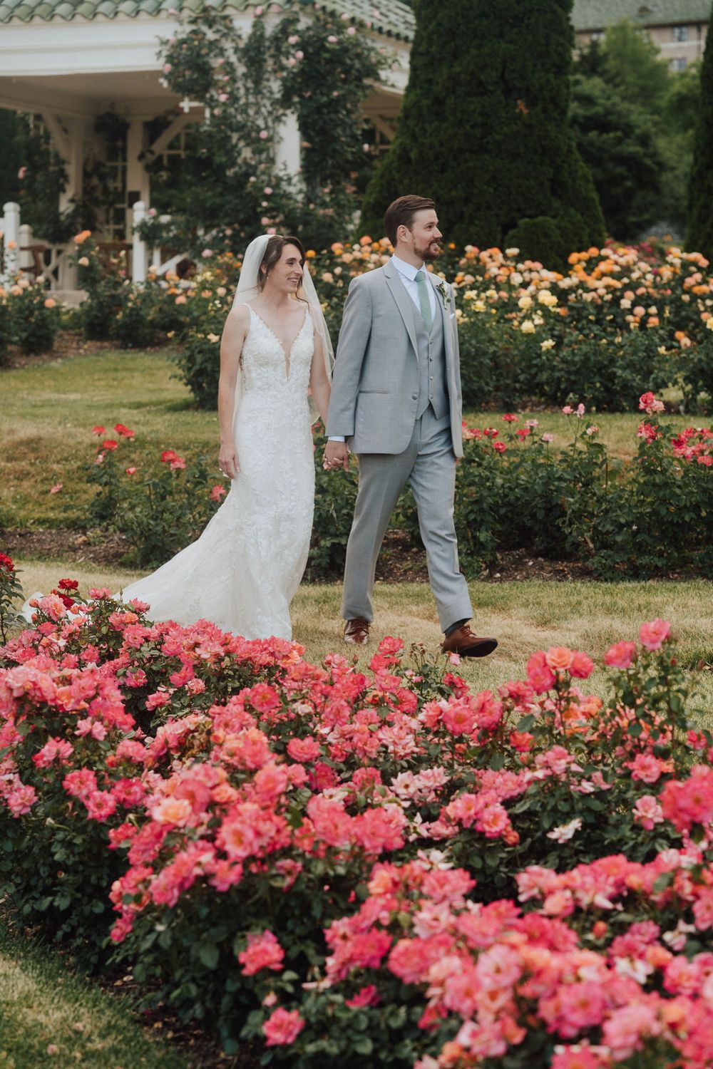 Bride and groom walk hand-in-hand through a vibrant rose garden in full bloom on their wedding day.