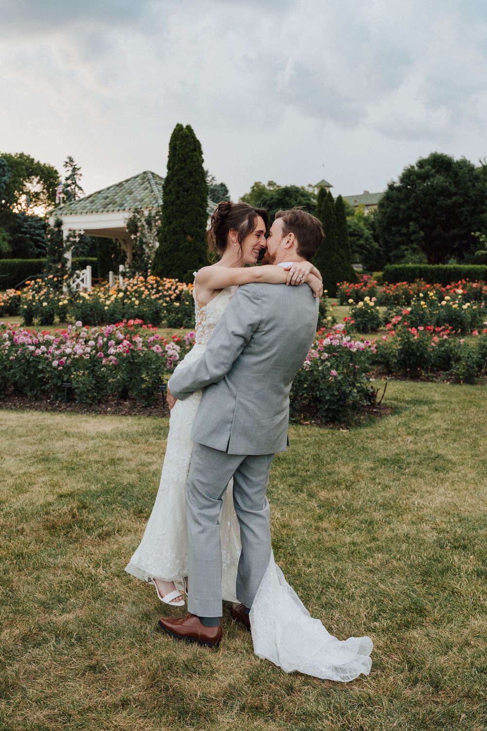 A bride and groom share a romantic embrace in a lush garden with colorful roses and greenery on their wedding day.