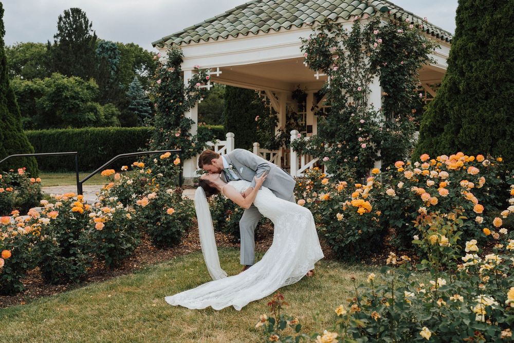 Bride and groom share a romantic dip kiss surrounded by colorful rose bushes near a white garden gazebo.