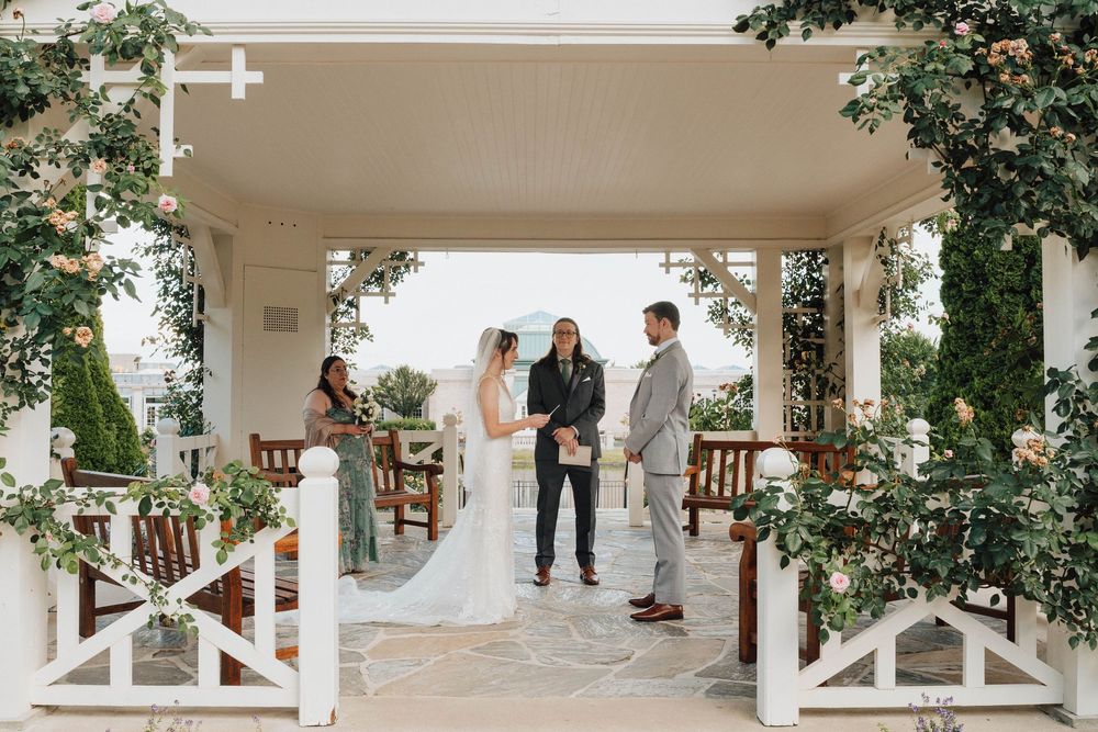 Couple exchanges vows under a white pergola adorned with greenery and florals at an elegant outdoor wedding ceremony.