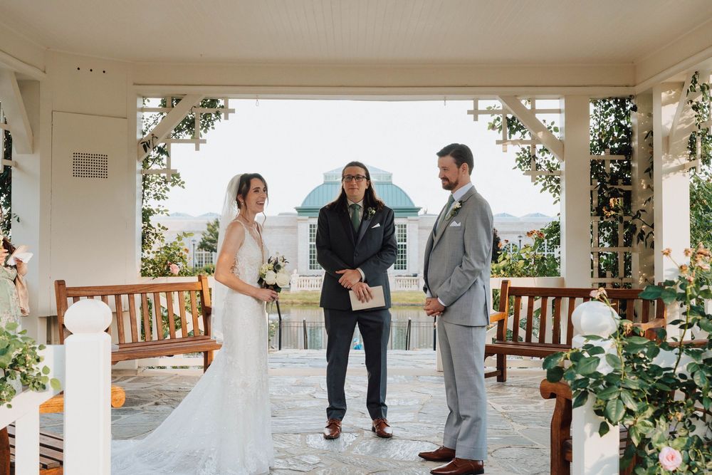Bride and groom exchange vows with officiant in elegant outdoor pavilion wedding ceremony surrounded by floral decor.