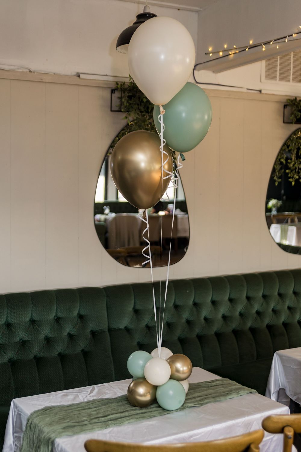 Elegant balloon centerpiece in gold, sage green, and white on a white-clothed table with green velvet seating.