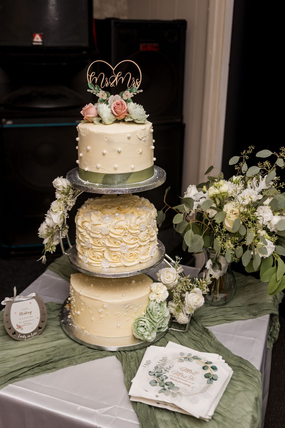 Three-tier white wedding cake with Mr & Mrs topper, rosette details, and floral decorations on a draped table.