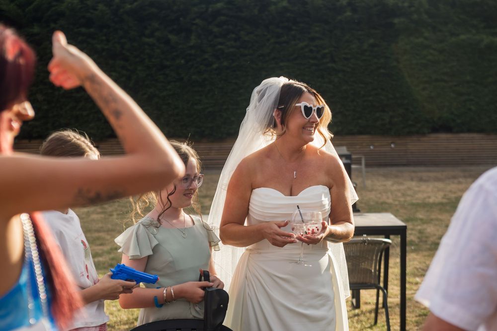 Bride in white strapless gown and veil wearing sunglasses smiles at outdoor wedding reception.