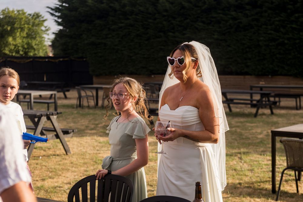 A bride in white strapless gown and veil wearing sunglasses stands with a young girl at outdoor wedding.