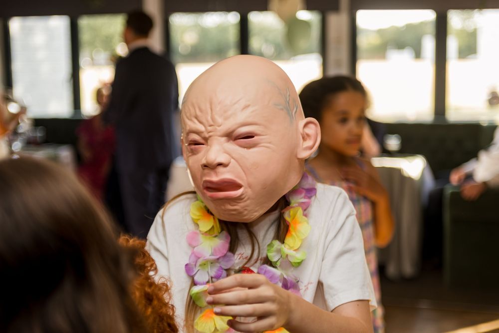 Person wearing a realistic crying baby mask with a floral lei at an indoor event.