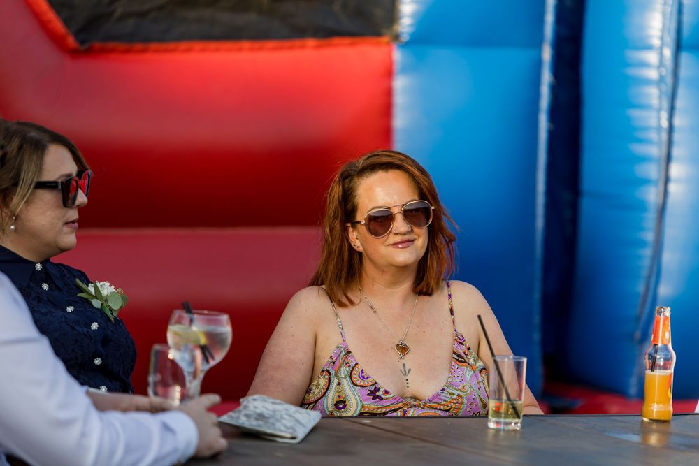 Two women in sunglasses relaxing at outdoor wedding reception near inflatable bouncy castle.