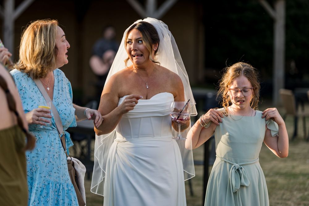 Bride in white gown and veil laughing outdoors with female guests at wedding.