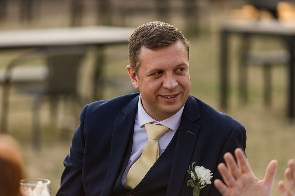 Smiling man in navy suit and gold tie with boutonniere seated at wedding.