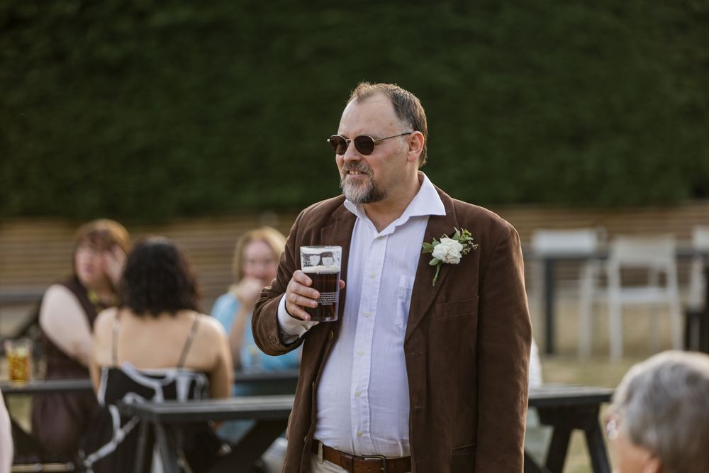Smartly dressed man in brown suit with boutonniere holding a pint of beer at outdoor wedding reception.