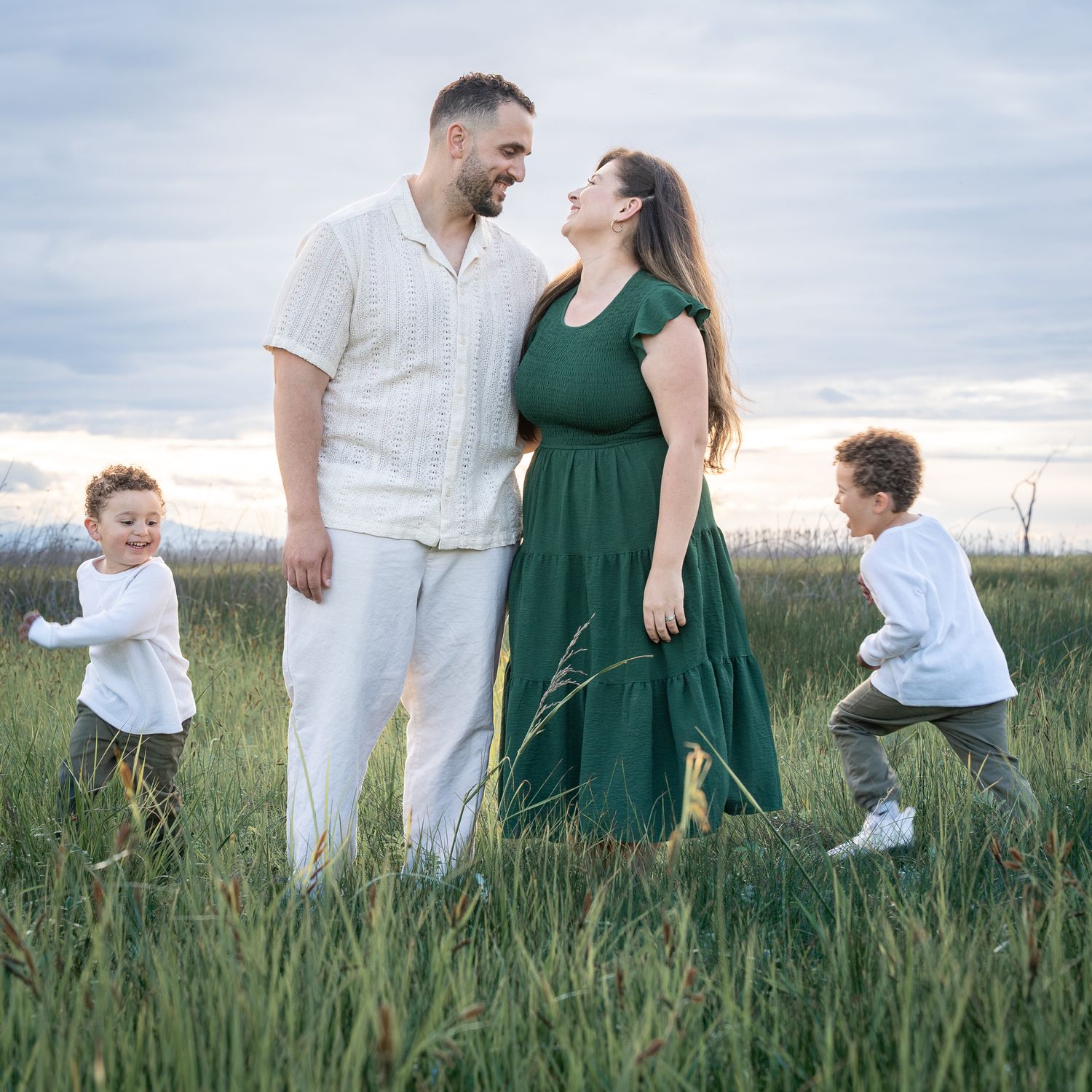 Parents smiling at each other in a grassy field at sunset while their two young boys run and play beside them.