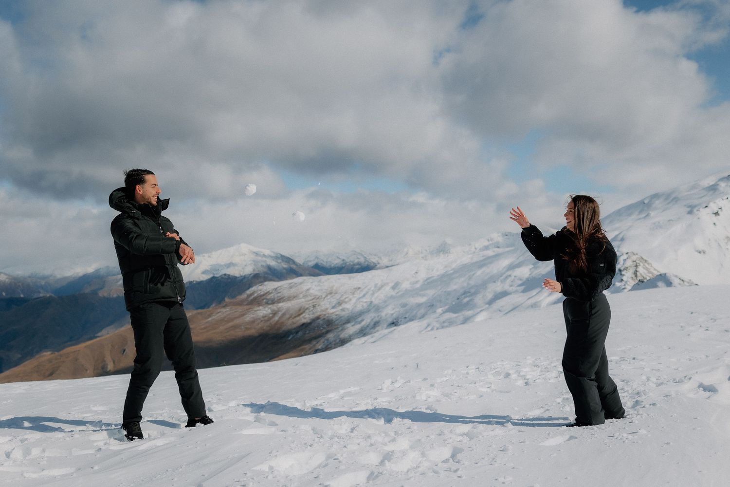 Man and woman in dark winter clothing stand on a snowy mountain ridge under a cloudy sky, with snow-covered peaks in the distance.