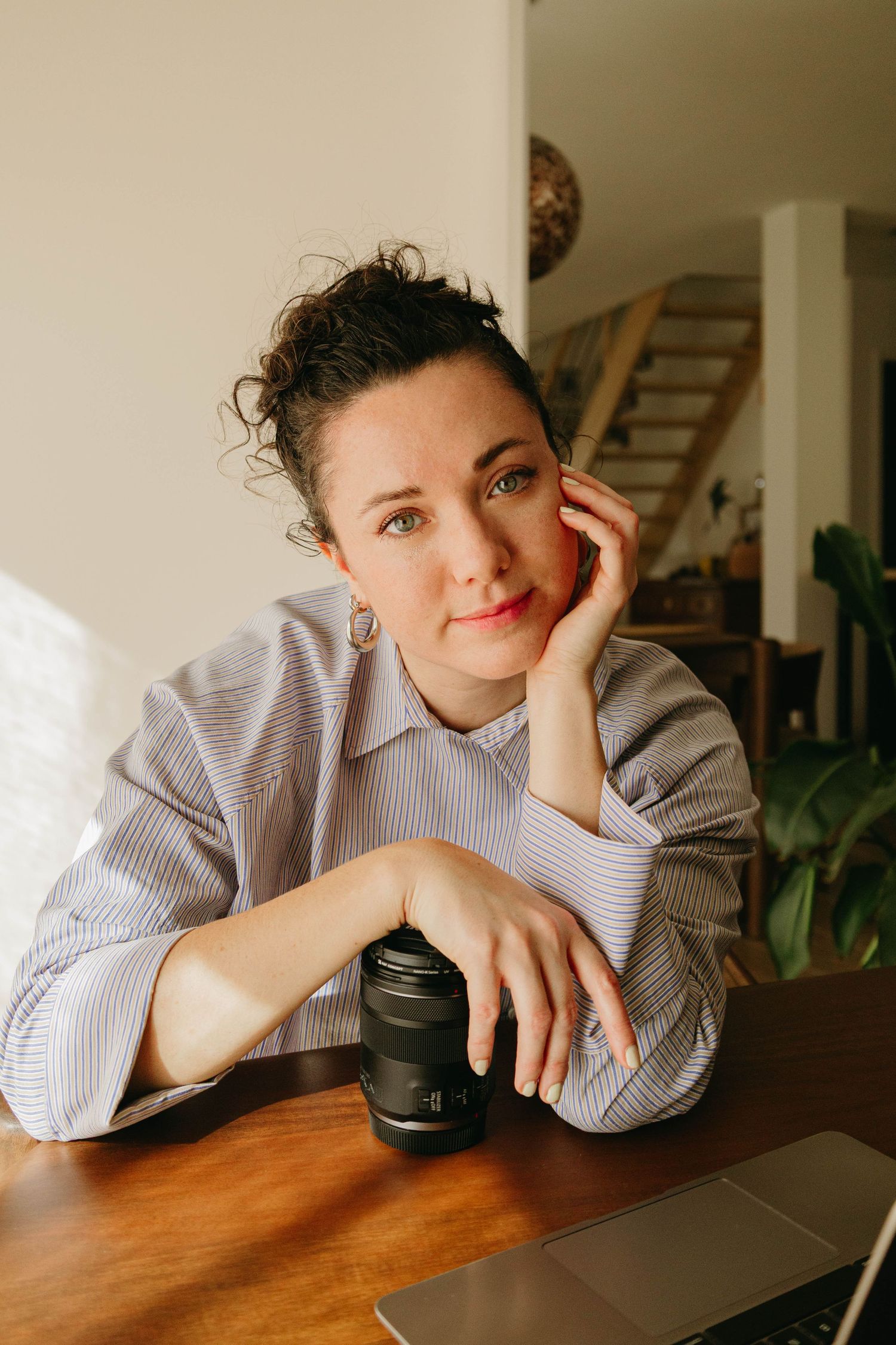 Woman in a striped button-up shirt rests her chin on her hand while holding a camera lens at a wooden table with a laptop.