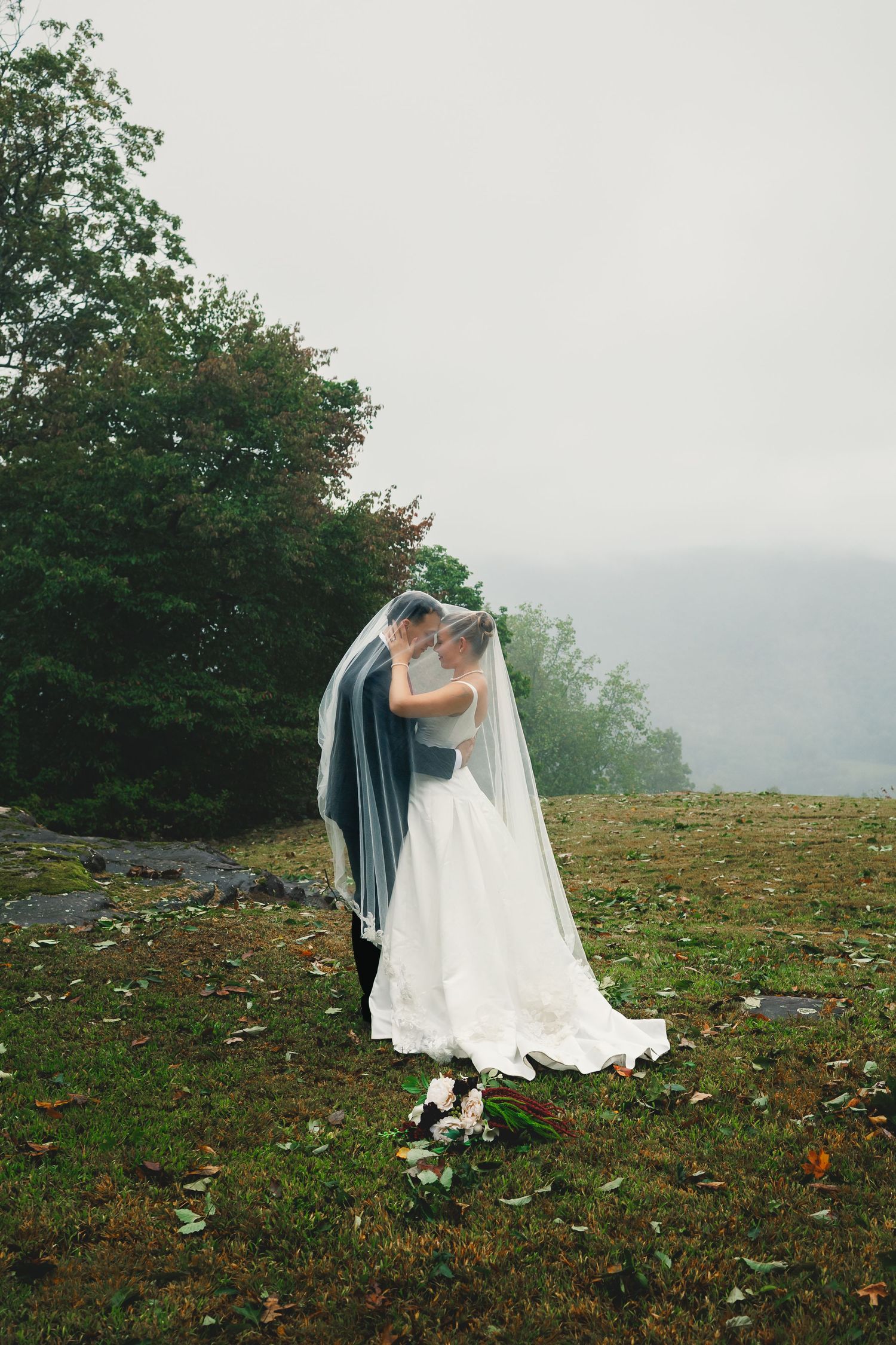 Bride and groom embrace under a sheer veil on a misty, grassy hilltop, with a bridal bouquet resting on the autumn leaf-covered ground.