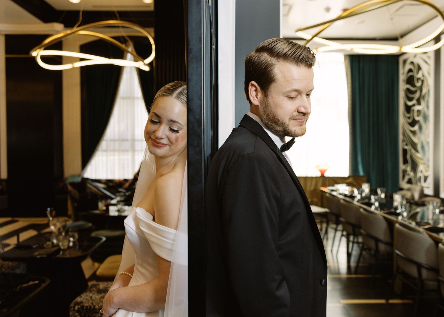 Bride in off-shoulder gown and groom in tuxedo lean back-to-back against a doorway in an elegant venue.