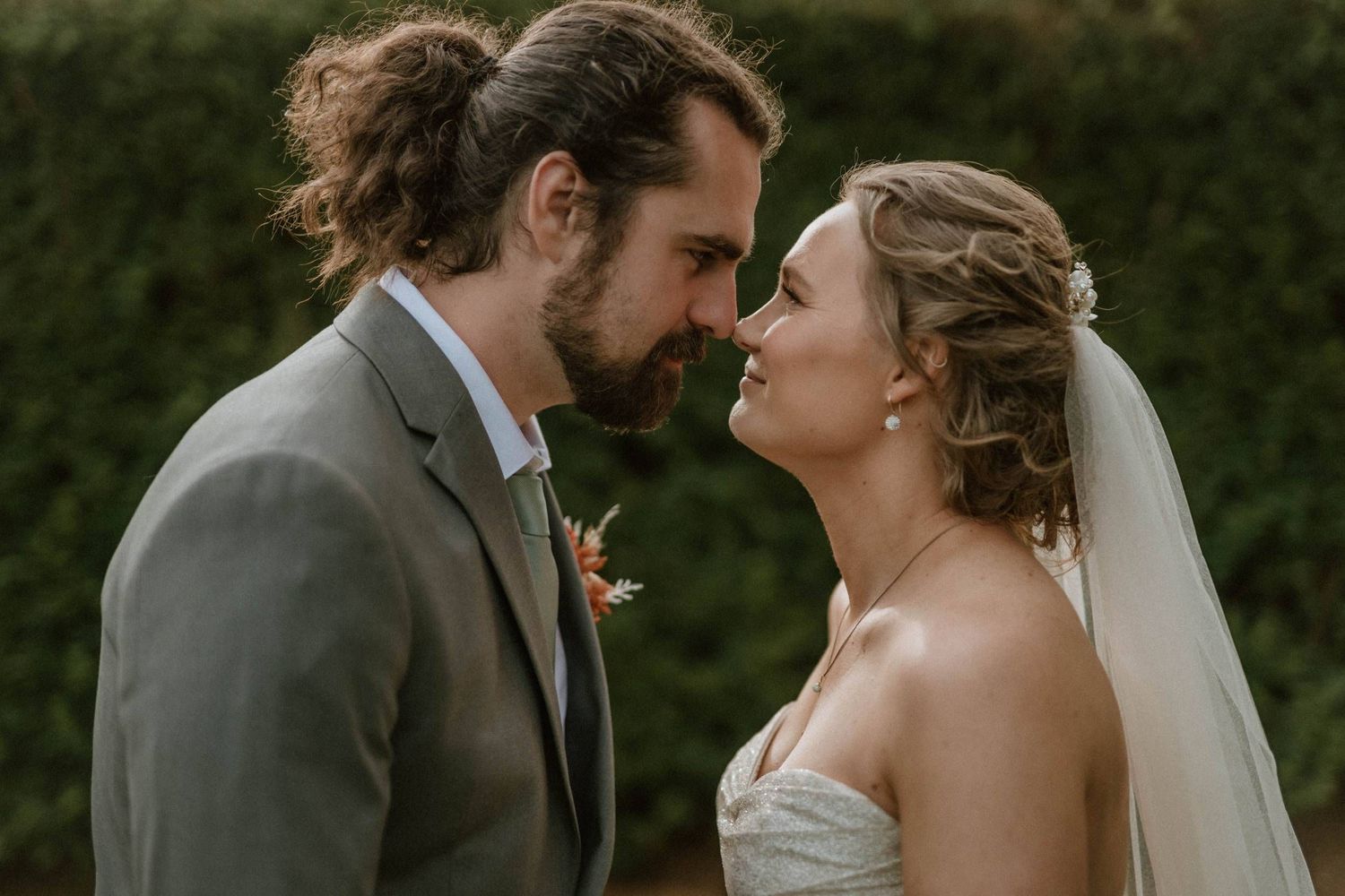 A bride and groom share an intimate moment, nearly kissing, dressed in wedding attire with a lush green hedge backdrop.