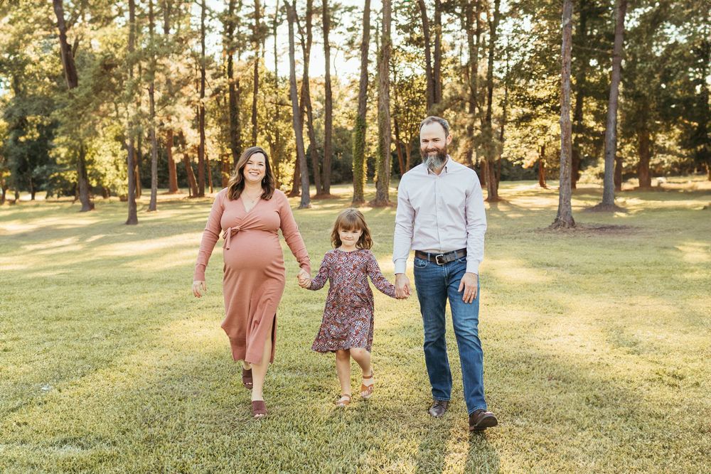 Pregnant woman and man walk with young girl through sunny park, holding hands in a family maternity photo session.