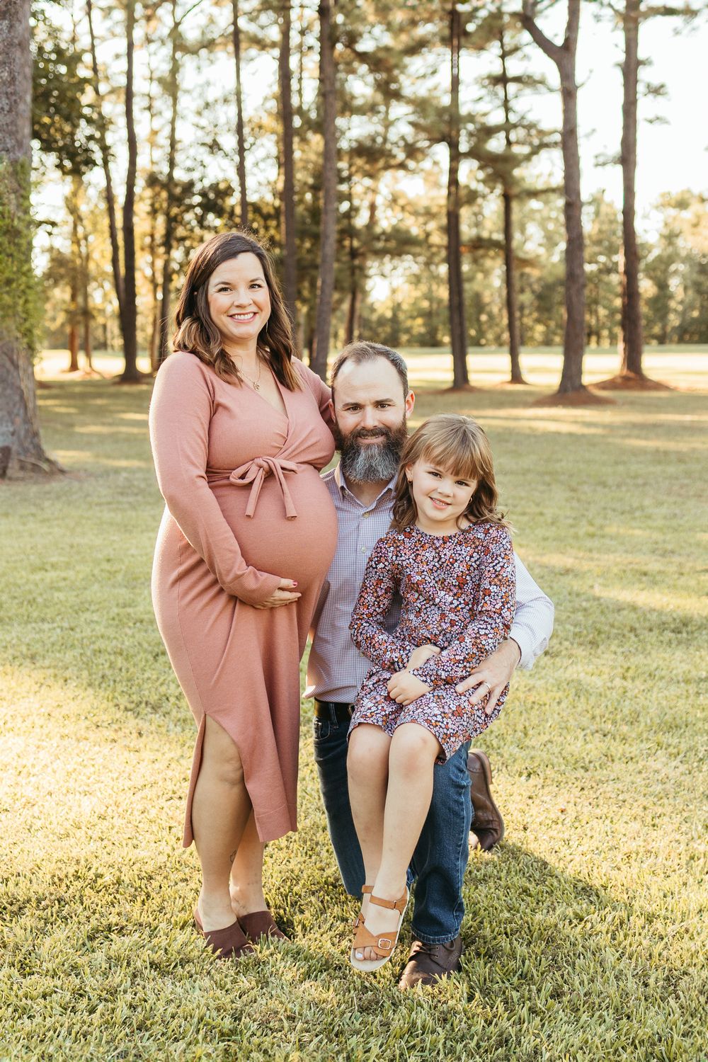 Pregnant woman in pink dress poses with bearded husband and young daughter outdoors among tall pine trees.