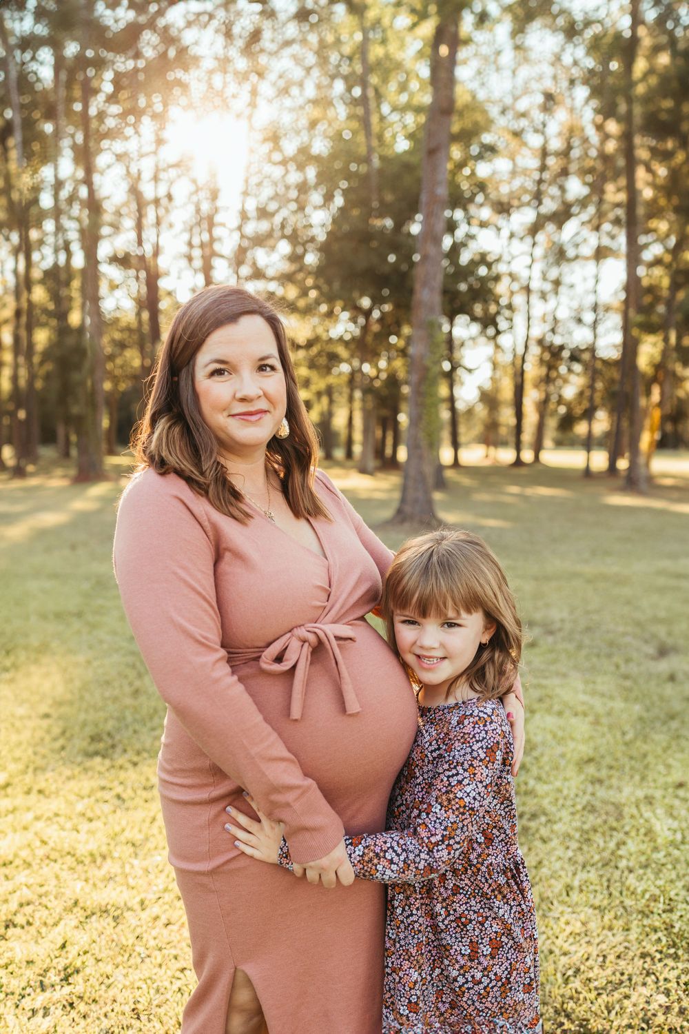 Pregnant woman in pink dress with young girl hugging her baby bump in a sunlit park setting.