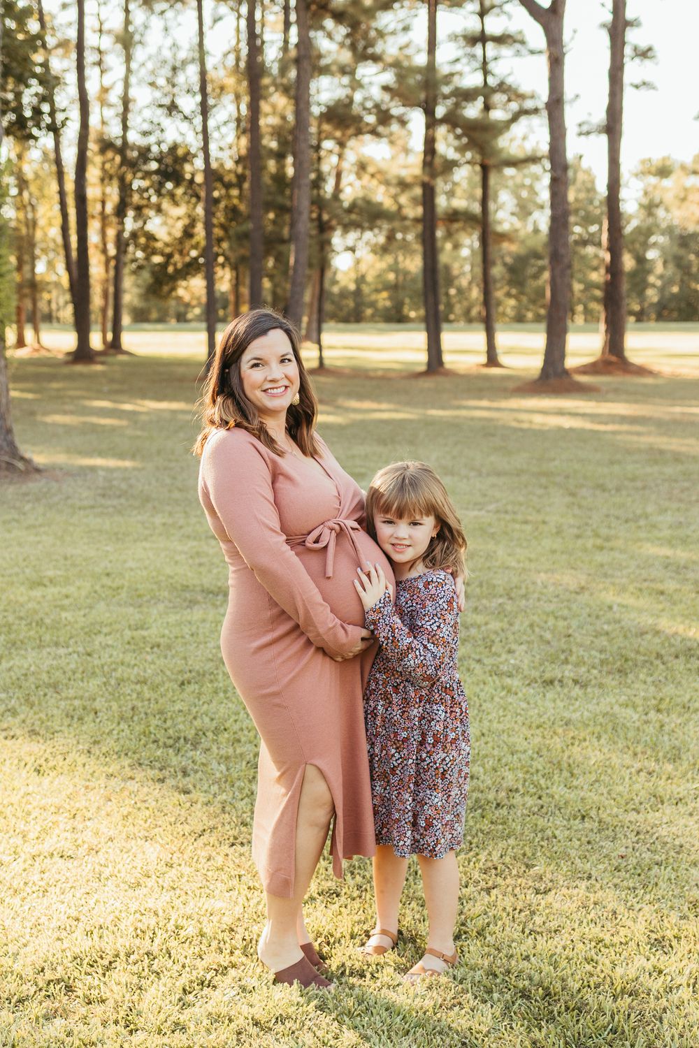Pregnant woman in pink dress hugging young daughter outdoors in sunny park during maternity photo session.