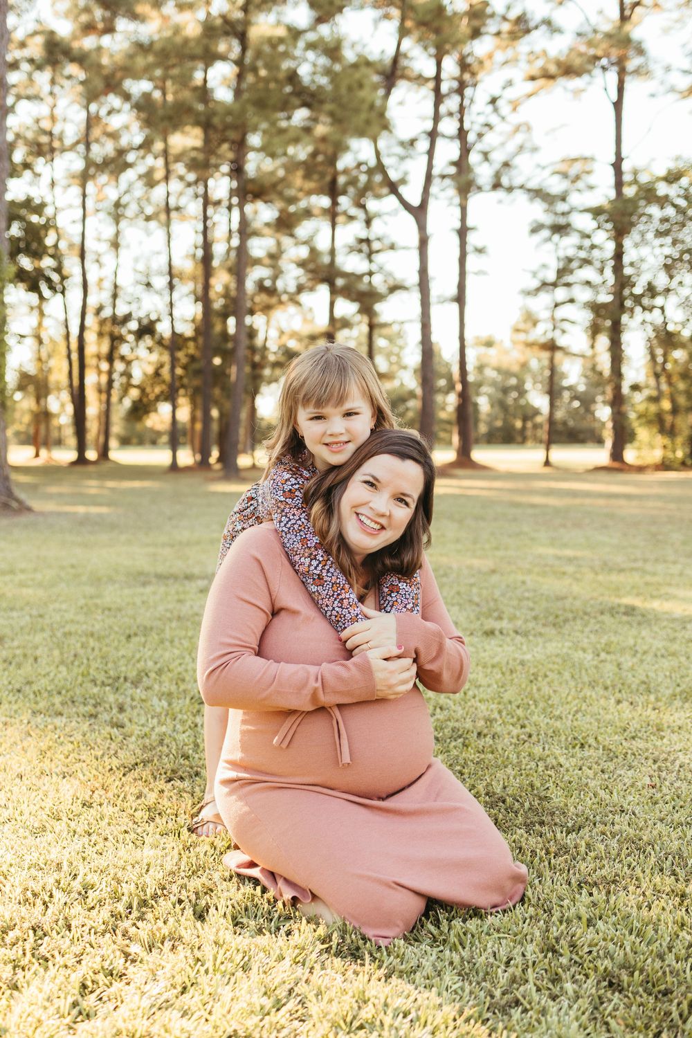 Pregnant mom in mauve dress kneeling on grass as young daughter hugs her from behind in sunny park setting.