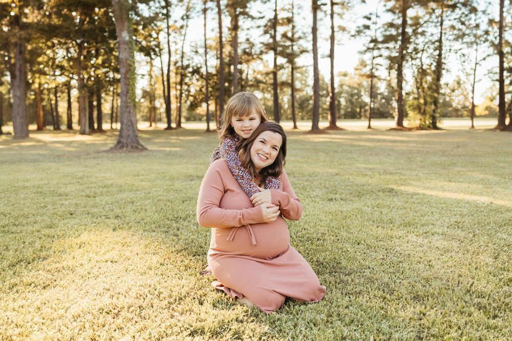 Pregnant woman in pink dress kneeling on grass while young girl hugs her from behind in sunny park.