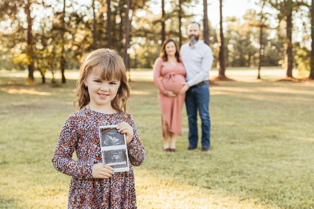 Young girl holds ultrasound photo while pregnant mother and father stand behind her in sunny park.