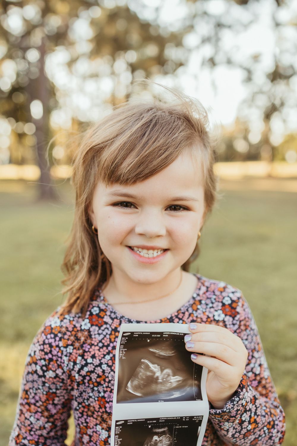 Smiling young girl in floral dress holding ultrasound photo outdoors in sunny park.