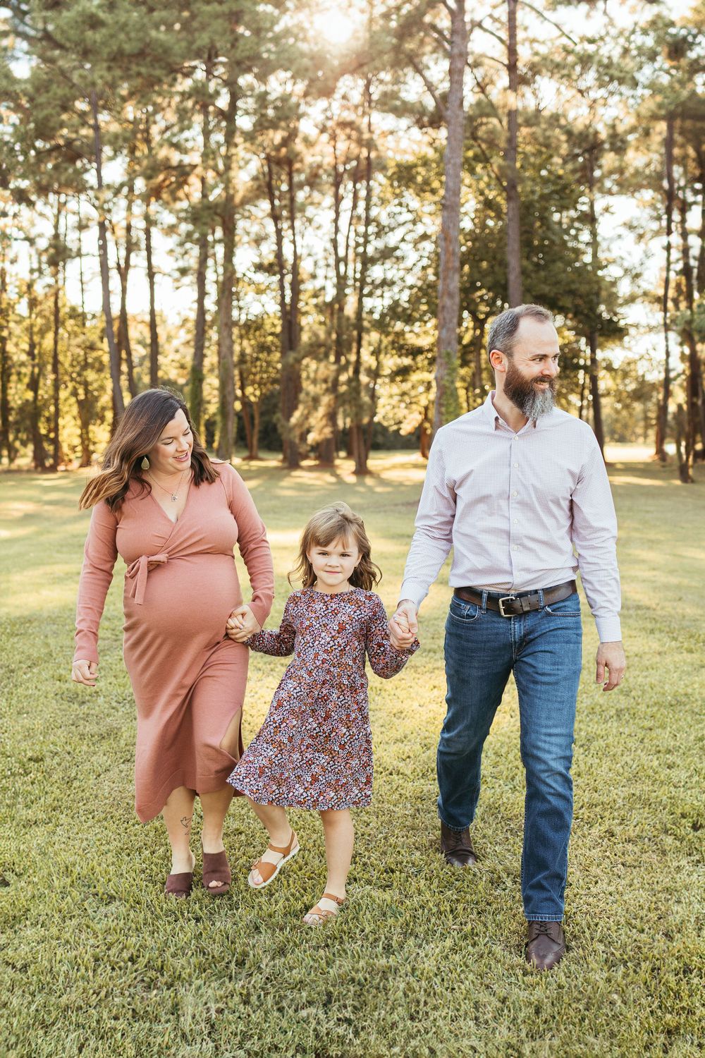 Pregnant woman in pink dress walks hand-in-hand with daughter and bearded husband in sunny park.
