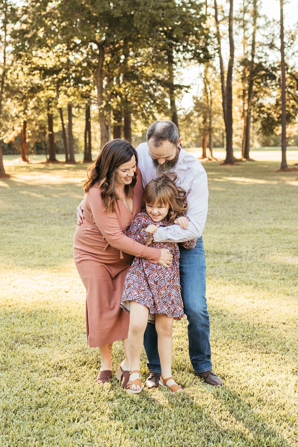 A happy family of three sharing a warm embrace outdoors in a sunlit park with tall trees in the background.