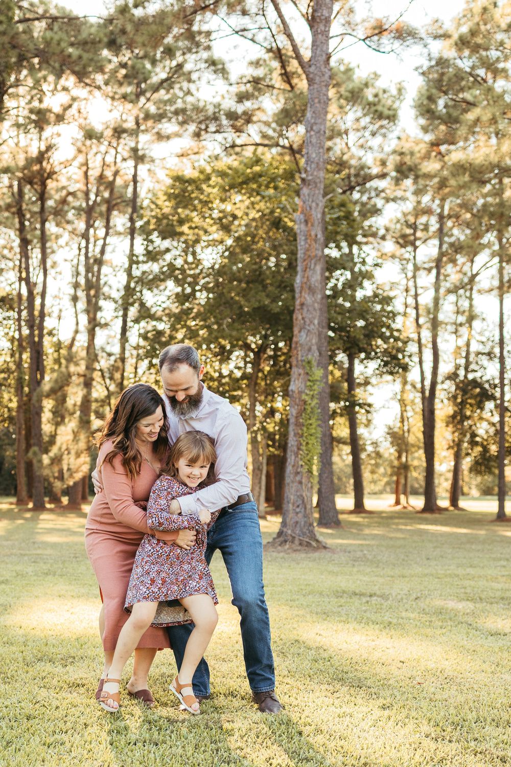 Family of three sharing a joyful moment in a sunlit park, parents embracing toddler daughter among tall pine trees.