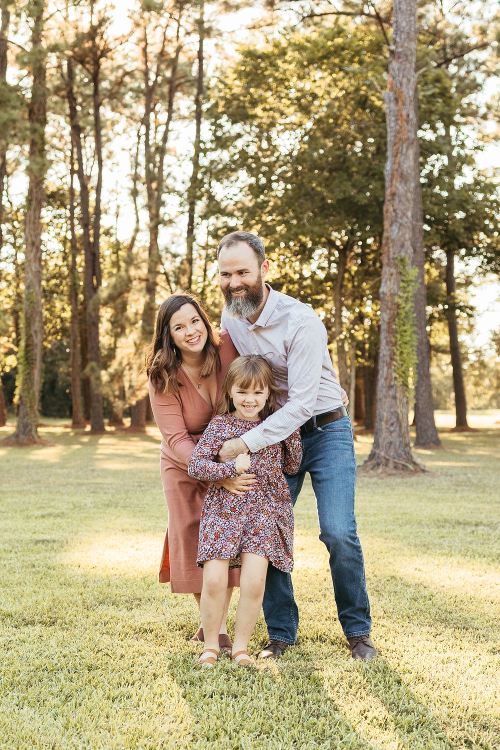 Family of three posing outdoors in golden hour light, parents embracing young daughter in floral dress on green lawn.