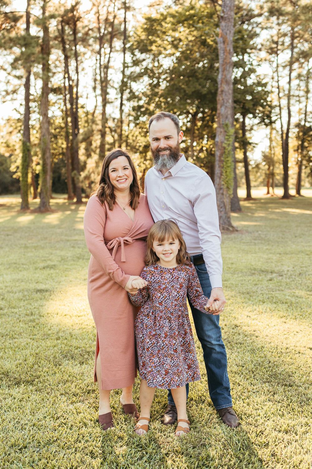 A smiling family of three poses outdoors in a sunny park, with a pregnant mother, bearded father, and young daughter.
