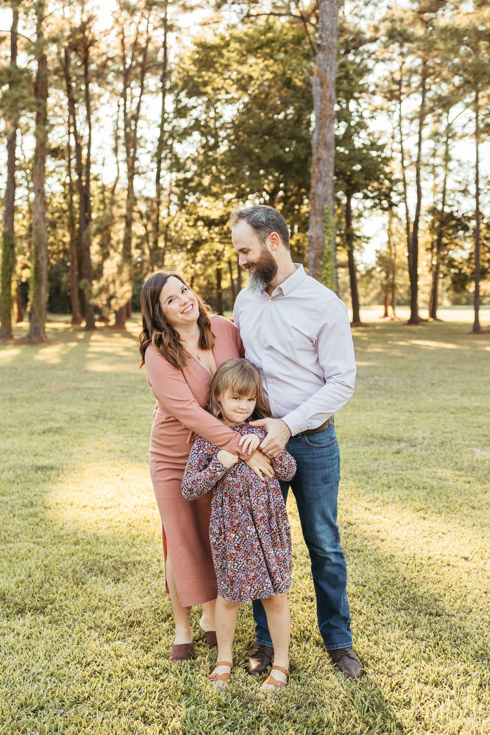 Family of three posing outdoors in golden hour light, mom in pink dress hugging daughter while dad smiles down at child.