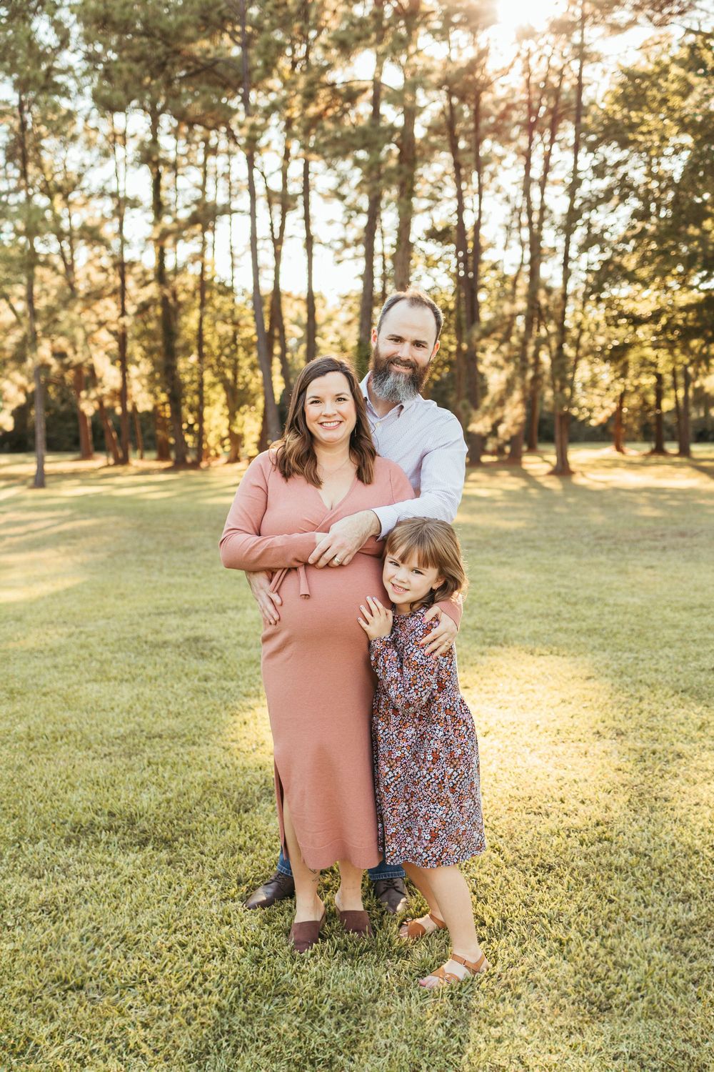 Pregnant woman in pink dress with husband and young daughter posing in sunlit park for maternity family portrait.