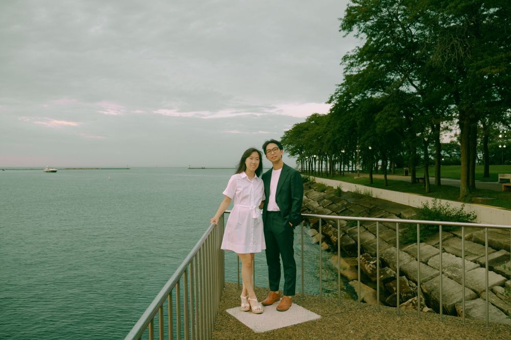 Couple posing together by a waterfront railing with lush green trees and cloudy sky in background.