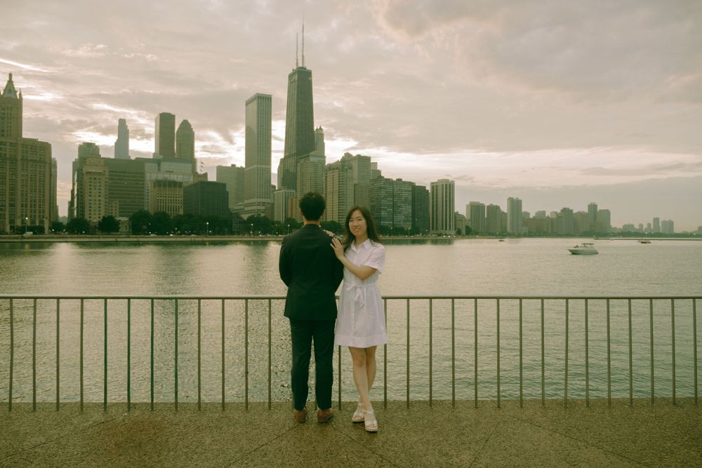 Couple standing by waterfront railing with Chicago skyline and cloudy sunset sky in background.