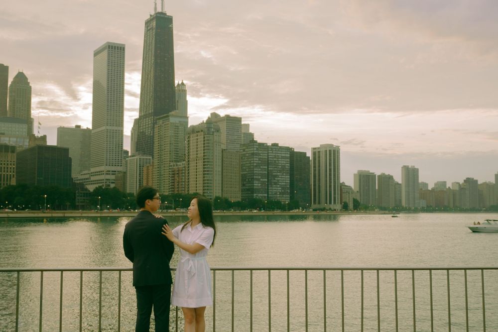 Couple standing by waterfront railing with Chicago skyline and John Hancock Center at sunset.
