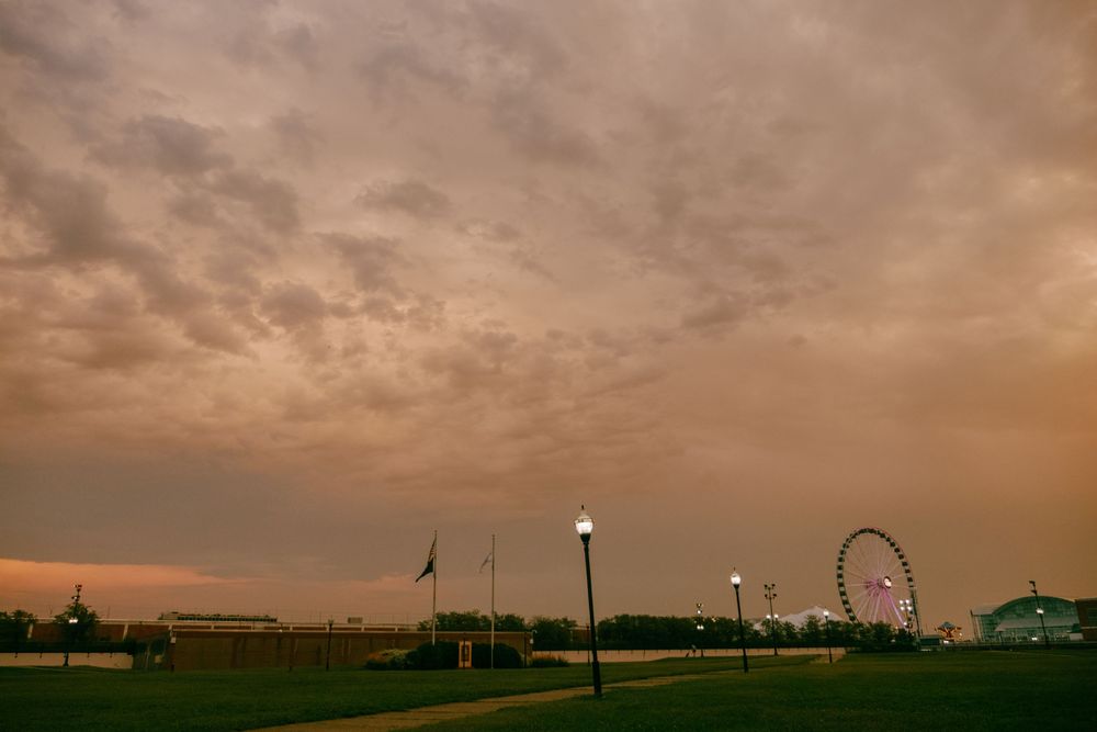 Dramatic sunset sky over a park with street lamps, flags, and a Ferris wheel silhouette on the horizon.