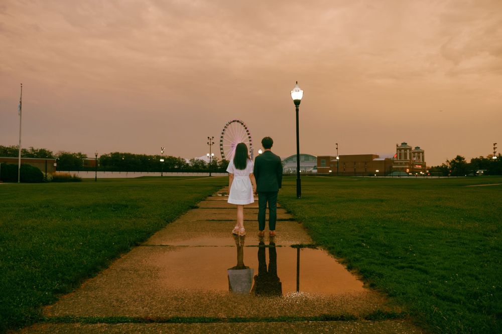 Couple standing on a pathway at dusk near a Ferris wheel, with a glowing street lamp and cloudy orange sky.