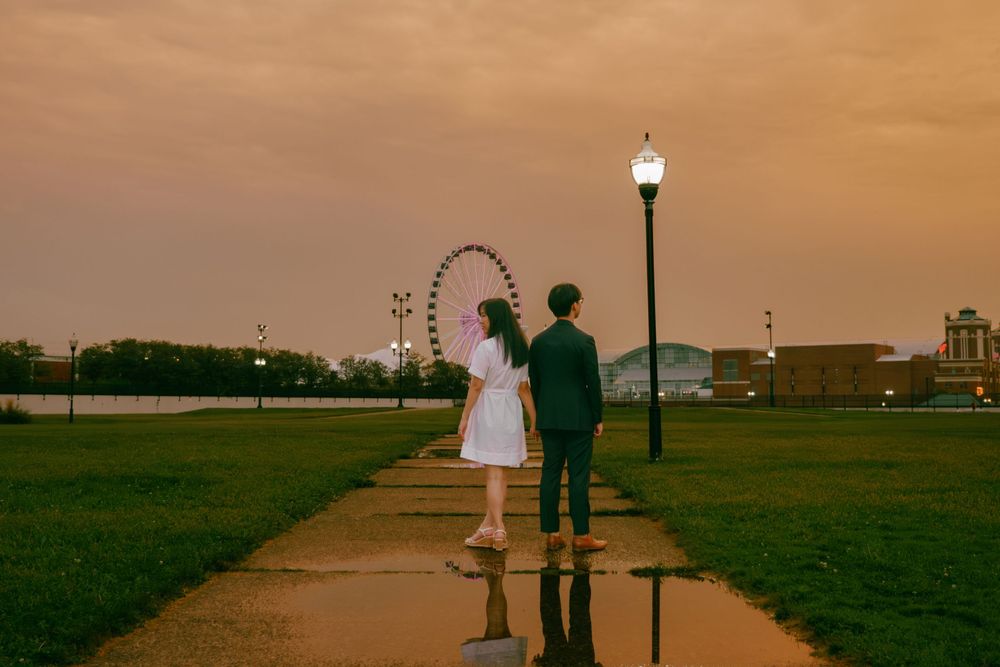 Couple walking hand-in-hand at dusk near a Ferris wheel and glowing street lamp on a reflective path.
