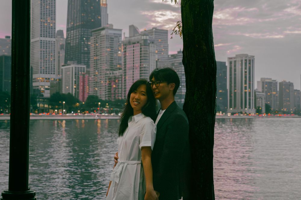 A young couple stands by a waterfront at dusk with a glowing city skyline reflecting on the water behind them.