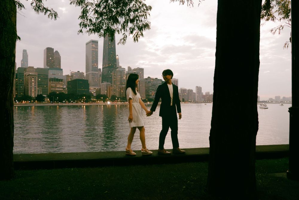 Couple holding hands walking by a city waterfront park with skyline views at dusk.
