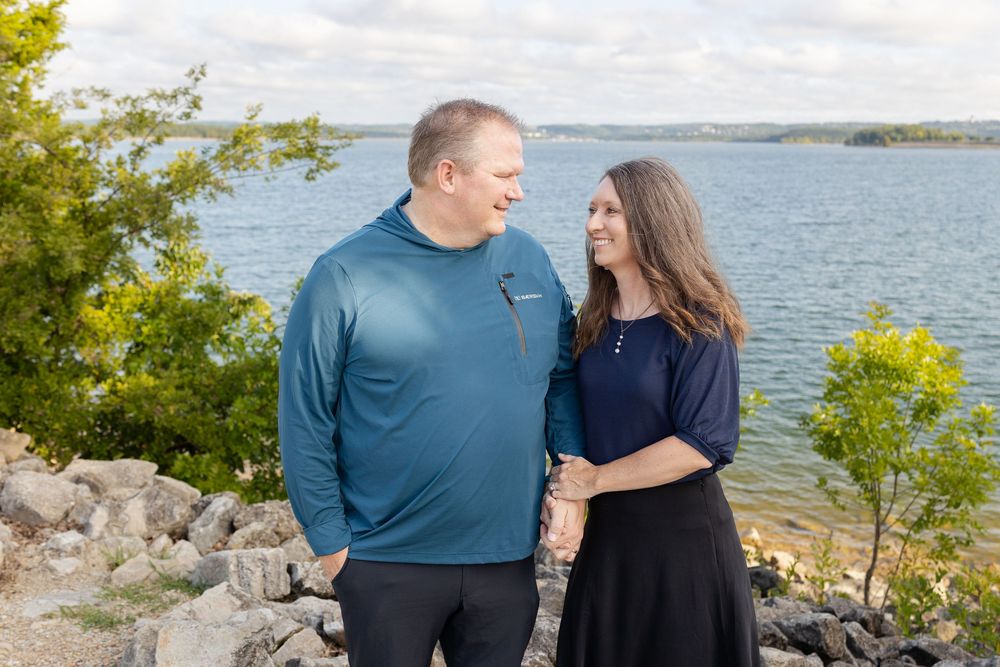 Couple smiling at each other near a rocky lakeside with trees and water in the background on a cloudy day.