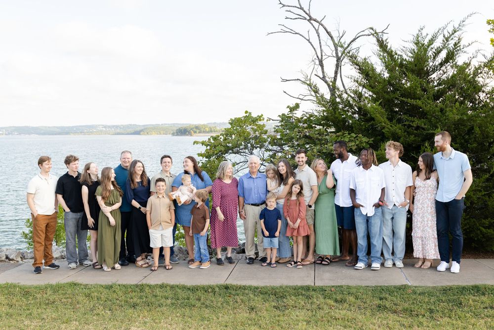 Large family group portrait outdoors by a lake, smiling together on a sunny day.